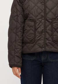 Quilted dark brown jacket with front snap buttons and two patch pockets worn with dark blue jeans.