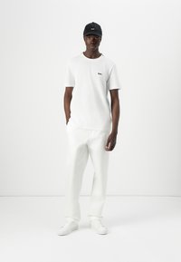 T-shirt en coton blanc avec un petit logo noir, associé à un pantalon blanc ample et des baskets blanches. Une casquette noire complète la tenue.