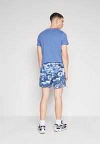 Camiseta de algodón azul con mangas cortas, combinada con bañador azul con estampado de palmeras y texto. Calcetines blancos y zapatillas.