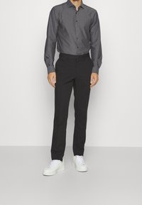Chemise à boutons gris foncé avec un motif subtil, associée à un pantalon noir et des baskets blanches, présentant une coupe ajustée et des manches longues.