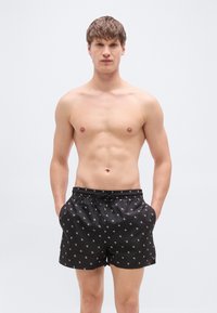 Μαύρα μαγιό βερμούδα με κορδόνι στη μέση, διακοσμημένα με μικρό λευκό λογότυπο και πλαϊνές τσέπες. Το ύφασμα φαίνεται λείο και ελαφρύ.