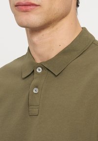 Prim-plan al feței inferioare și gâtului unui bărbat care poartă un tricou polo verde măsliniu cu doi nasturi albi vizibili.