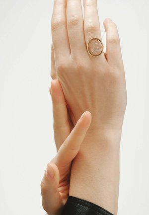 Mano con anillo ovalado de oro grabado con una pequeña flor, sostenida suavemente por una segunda mano contra un fondo claro y liso.