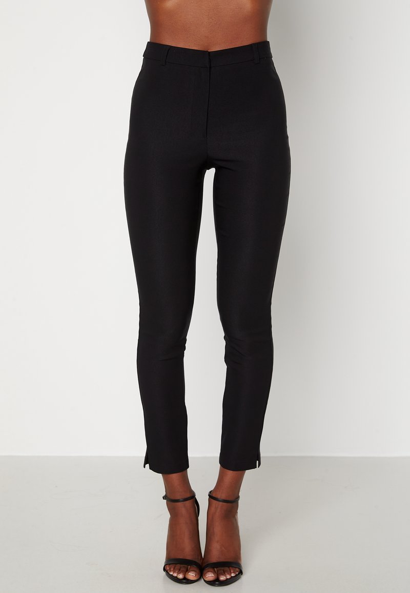 Pantalons noirs ajustés avec une texture lisse, taille haute et fentes latérales au bas. Associés à des escarpins noirs à brides.