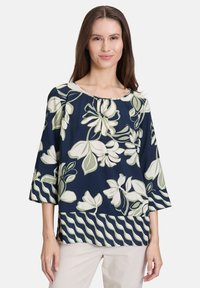 Granatowa bluzka w kwiaty z zielonymi i białymi wzorami, z okrągłym dekoltem i rękawami o długości trzech czwartych, wykonana z lekkiego materiału.