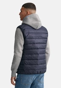 Chaleco de plumas navy con textura de acolchado horizontal, usado sobre una sudadera gris con capucha, con un cuello alto y sin cierres visibles.