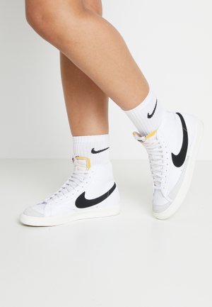 Legs wearing white Nike high-top sneakers with black swoosh logo and white crew socks featuring small black Nike logo on a white floor.
