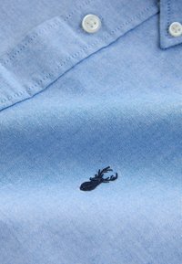 Chemise bleue claire à boutons avec un tissu texturé et tissé. Présente un petit logo brodé en forme de cerf noir près du bas.