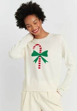 Pull en tricot crème avec un motif de canne en sucre rayée rouge et blanche, orné d'un nœud vert. Texture douce, encolure arrondie et poignets côtelés.