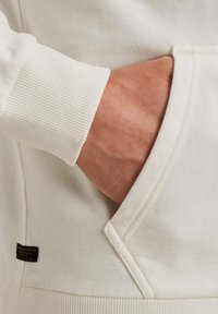 Krémová mikina s vreckom, rebrovanými manžetami a malou nášivkou po boku. Mäkká textúra a neformálny dizajn.