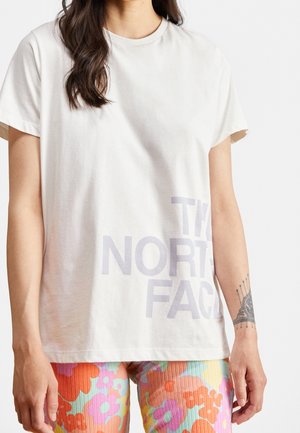 Osoba nosząca białą koszulkę z częściowo widocznym tekstem "The North Face" oraz kolorowe spodnie w kwiatowy wzór.