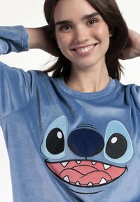 Sudadera de felpa azul con un gran rostro del personaje Stitch bordado, con ojos prominentes y una boca sonriente. Mangas largas, textura suave.