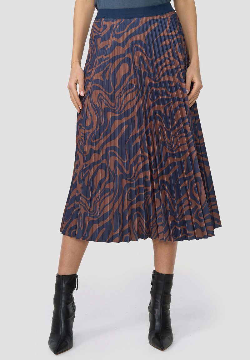 Žena v plisé sukni midi s hnědým a navy abstraktním vzorem, s navy pasem, černými špičatými kotníkovými botami, stojící s uvolněnýma rukama.
