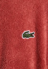Tissu éponge rouge avec une surface texturée et un logo de crocodile vert bordé de blanc brodé sur le côté gauche.
