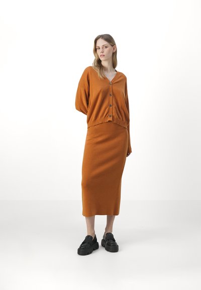 Cardigan en tricot orange avec de grands boutons, assorti à une jupe midi. L'ensemble présente une coupe décontractée et une texture douce, accompagné de chaussures noires.
