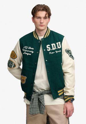 Jeune homme porte une veste universitaire verte et crème avec des écussons et des inscriptions, un pantalon beige et une chemise à carreaux nouée à la taille.