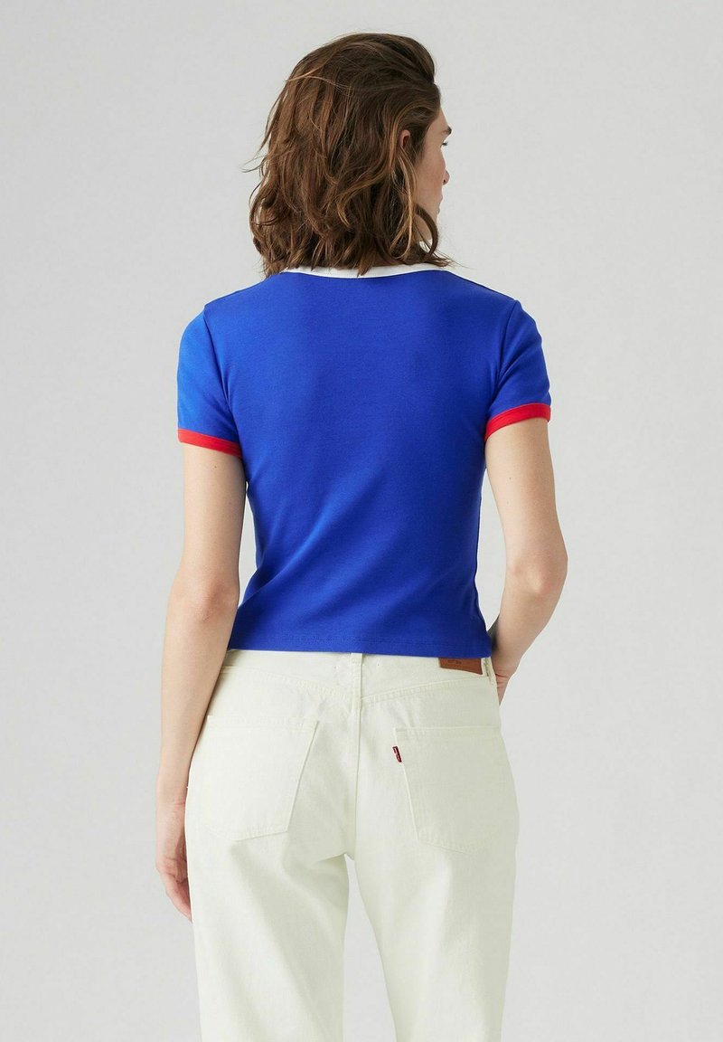 Camiseta recortada azul con acentos rojos y blancos en las mangas y el escote, combinada con unos jeans de color claro. Tejido suave, diseño casual.