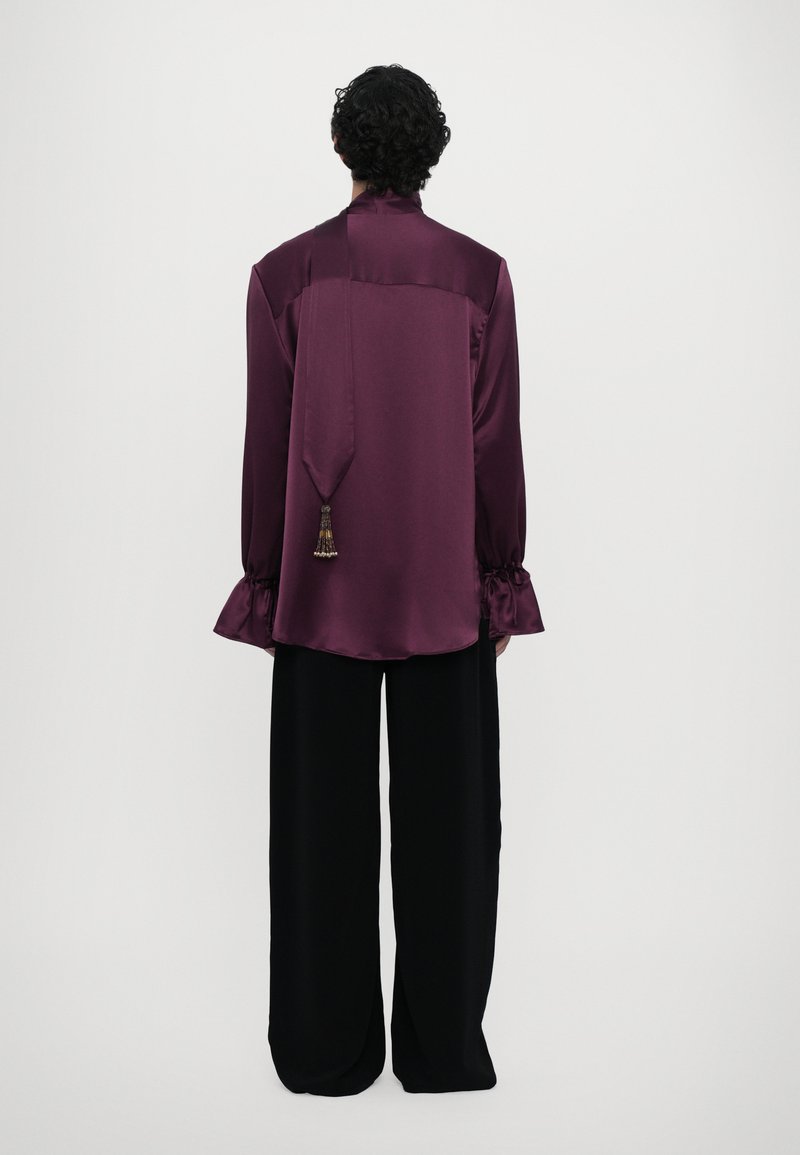 Maroon satin blouse with a high collar, long sleeves, and ruffled cuffs, featuring a back tie detail with a decorative tassel. Paired with black wide-leg pants.