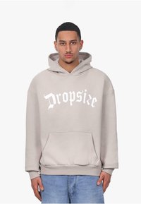 Beige übergroßer Hoodie mit einer vorderen Kängurutasche und Kapuze mit Kordelzug. Auf der Brust ist der weiße Gothic-Text "Dropsie" zu sehen.