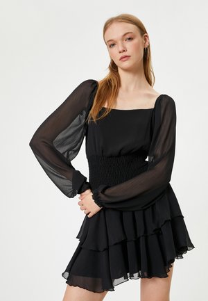 Vestido negro con mangas largas transparentes y abullonadas, un cuerpo fruncido y una falda de volantes en capas. Tela texturizada con un diseño fluido.