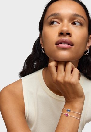 Bracelets en chaîne argentée ornés de charms floraux bleus et roses. Le mannequin porte un top sans manches crème, accompagné de boucles d'oreilles simples en forme de créole.