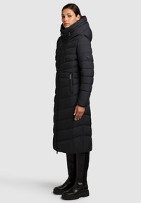 Svart pufferjacka med en lång design, hög krage och huva. Har horisontell quiltning och fickor med dragkedja. Matchas med knähöga svarta stövlar.