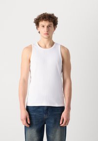 Débardeur blanc à côtes avec un col rond, coupe ajustée et sans manches, associé à un jeans en denim bleu. Texture lisse et design simple.