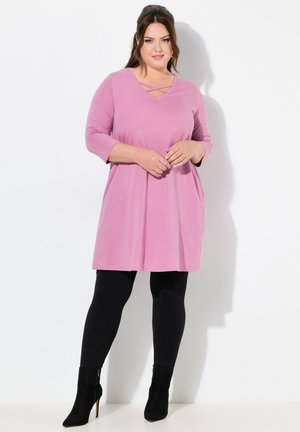Rosa Tunika-Kleid mit V-Ausschnitt und Kreuzdetail, langärmlig, lockere Passform, mit seitlichen Taschen, kombiniert mit schwarzen Leggings und Absatzstiefeln.