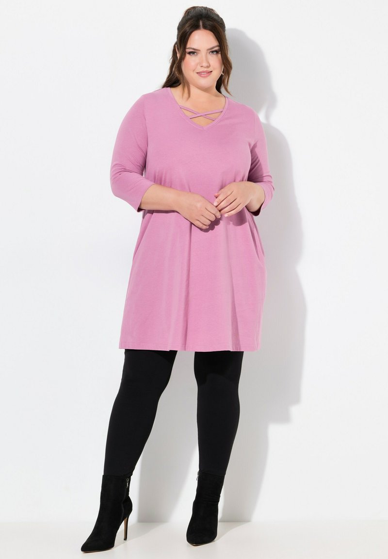 Rosa Tunika-Kleid mit V-Ausschnitt und Kreuzdetail, langärmlig, lockere Passform, mit seitlichen Taschen, kombiniert mit schwarzen Leggings und Absatzstiefeln.