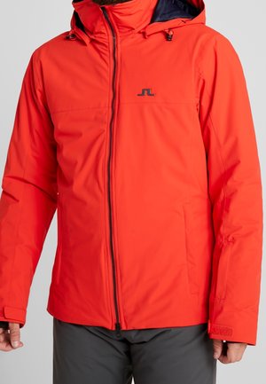 Hombre con chaqueta naranja brillante con cremallera, capucha y correas ajustables en las muñecas, combinado con pantalones gris oscuro.