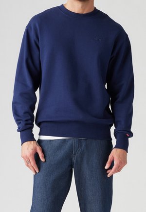 Mann trägt ein schlichtes marineblaues Levi's Sweatshirt über einem weißen Hemd und blauen Jeans, steht vor einem neutralen Hintergrund.