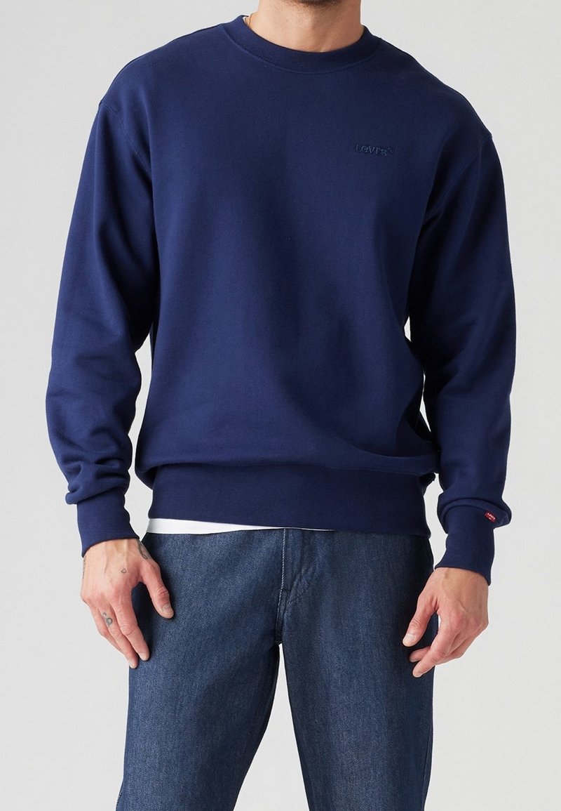 Homme portant un sweat-shirt uni Levi's bleu marine par-dessus une chemise blanche et un jean bleu, debout devant un fond neutre.