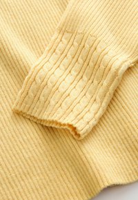 Nahaufnahme eines gelben gestrickten Pulloverarms mit Ripp- und Zopfmuster auf weichem Stoff.