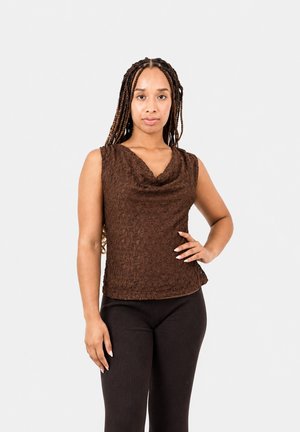 Femme aux longues tresses portant un haut marron texturé sans manches et un pantalon foncé côtelé, debout une main sur la hanche devant un fond clair.
