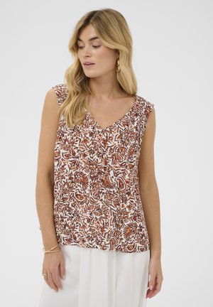 Naine, kellel on pikad blondid juuksed, seljas varrukateta pruun-valge lillemustriga pluus ja valge seelik, vaatab alla ja vasakule.