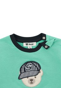 Camiseta de algodón verde claro con un oso con una gorra a rayas bordada en el frente. Cuello ribeteado negro con cierre a presión.