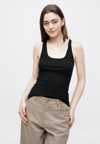 Mlada žena s dugom smeđom kosom, odjevena u crni top bez rukava i bež hlače, stoji ispred jednobojne bijele pozadine.