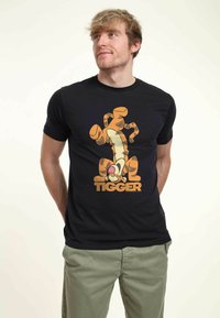 Svart t-shirt med en orange Tigger-karaktärsgrafik och ordet "TIGGER" i fetstil. Korta ärmar, klassisk passform.