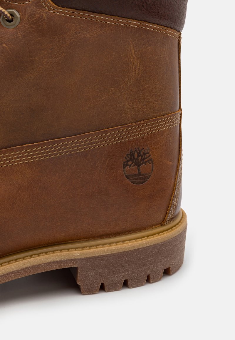 Botte en cuir marron avec une texture rugueuse, des accents marron foncé contrastants, des détails cousus et un logo d'arbre embossé sur le côté.