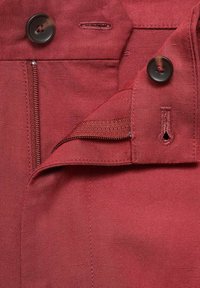 Pantalon en tissu rouge avec fermeture éclair et doubles accents de bouton. Il présente une texture lisse et un subtil détail de couture le long de la taille.