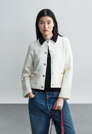 Žena v bielej zapínacej bunde cez čiernu košeľu s golierom, modrých džínsoch a drží červený remienok s kovovými očkami.