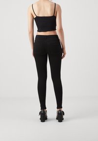 Femme debout, dos à la caméra, portant un débardeur noir court, un jean skinny noir taille haute et des bottines noires à talons.