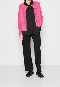 Ljusrosa stickad cardigan med svarta knappar, bärs över en svart skjorta med knappar och lösa svarta cargobyxor. Svarta klumpiga skor.