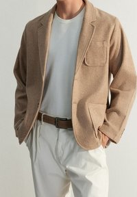 Blazer di lana marrone con una tasca sul petto, indossato sopra una camicia grigia chiara. Abbinato a pantaloni color crema e una cintura marrone.