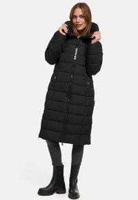 Manteau matelassé noir avec une silhouette longue, fermeture éclair, capuche et texture matelassée. Associé à un jean gris et des bottines noires.
