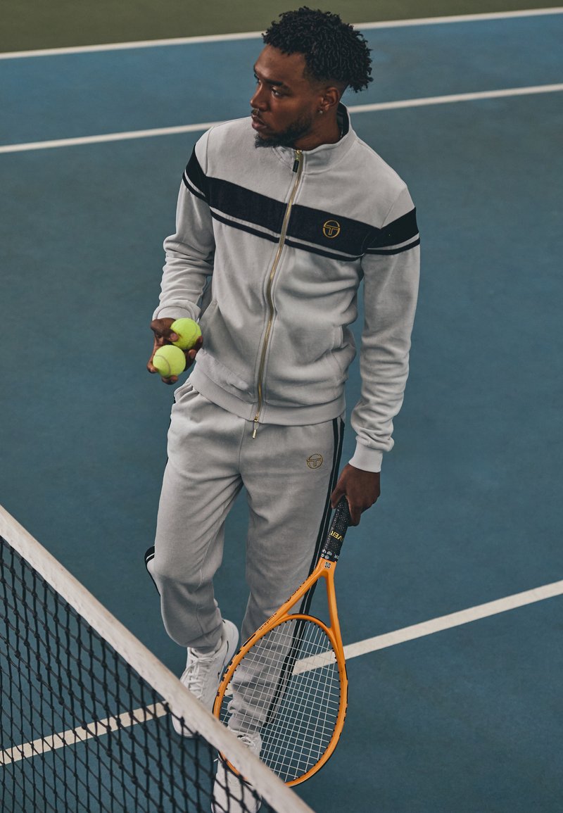 Grå treningsdrakt med svarte striper, som holder to grønne tennisballer og et oransje racket, står på en blå tennisbane.