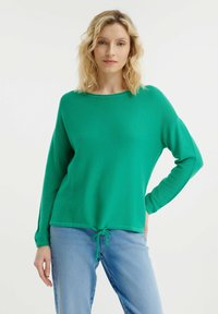 Grüner Strickpullover mit langen Ärmeln und rundem Halsausschnitt. Verfügt über einen Kordelzug am Saum und eine lockere Passform. Kombiniert mit hellblauen Jeans.