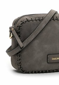Sac bandoulière en daim gris avec une fermeture éclair, orné de coutures décoratives le long des bords, une bandoulière plate et une plaque logo dorée.