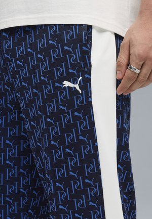 Pantalones deportivos azul marino con un patrón repetitivo de logotipos blancos y azules, con una franja lateral blanca contrastante y textura fina.