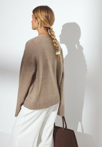 Brun stickad tröja med en avslappnad passform, ribbade ärmslut och nederkant, kombinerad med vita byxor och en mörkbrun toteväska. Lång flätad frisyr.